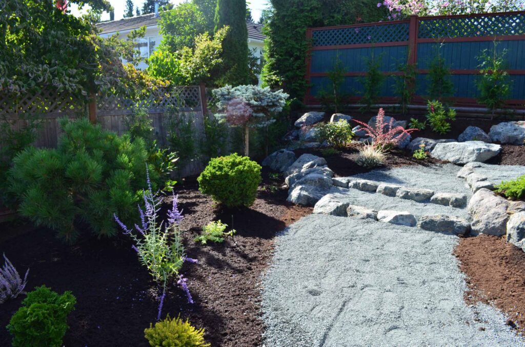 DSC_1075-scaled Landscaping example: A shrub/flower bed is accessible through a pebbled pathway. Two steps are outlined with larger stones.